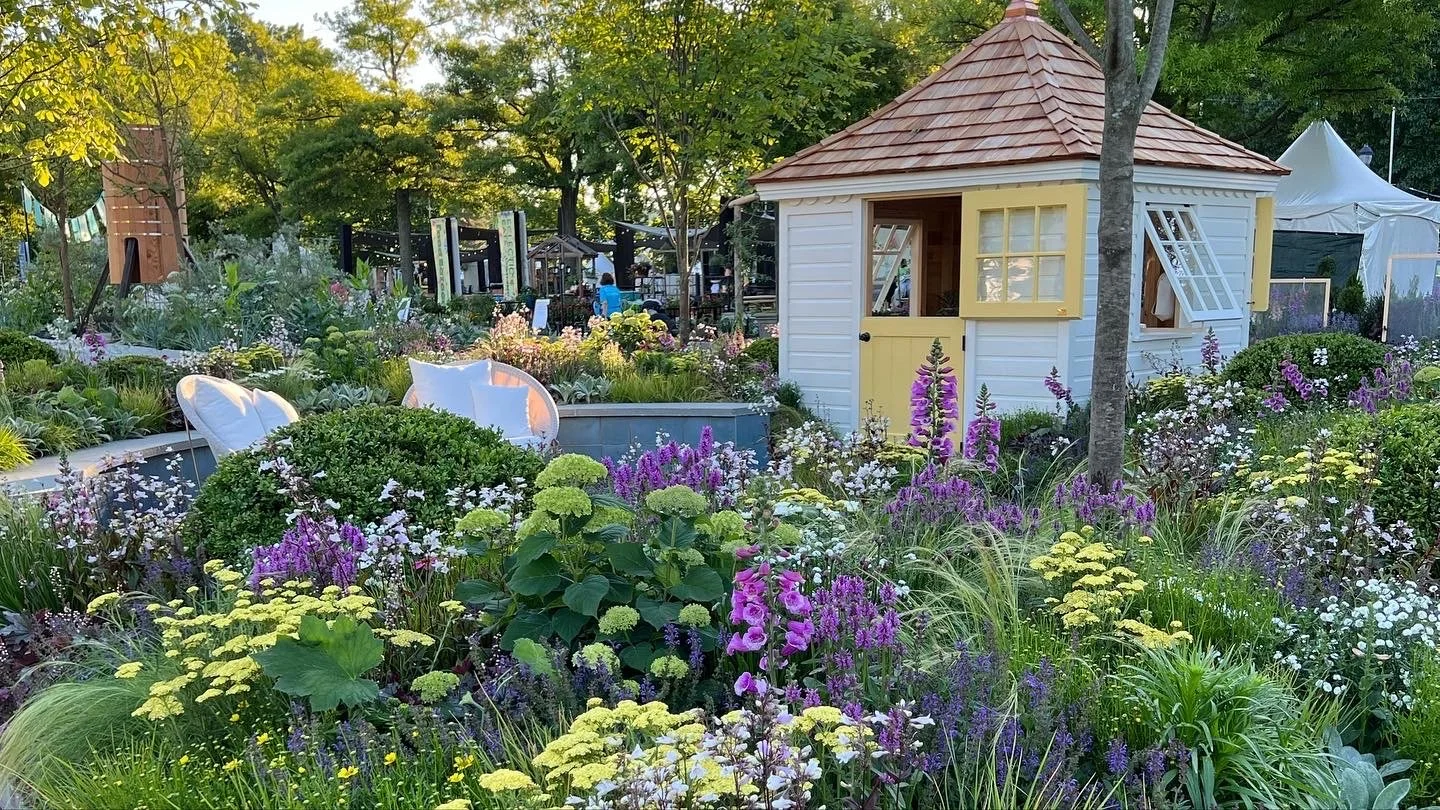 A lush garden with a variety of colorful flowers, a small white and yellow garden shed, and surrounding trees.