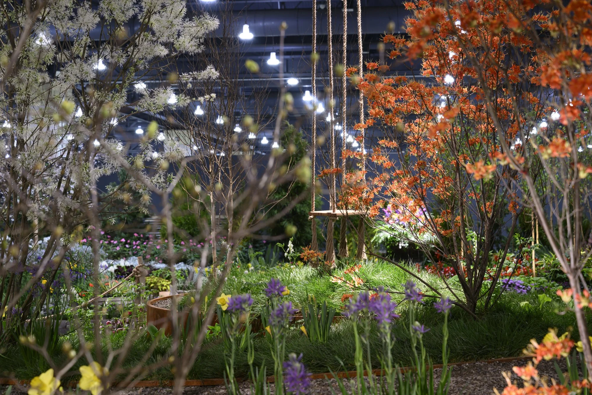 Indoor garden with blooming trees, colorful flowers, a wooden swing, and decorative plants under artificial lights.