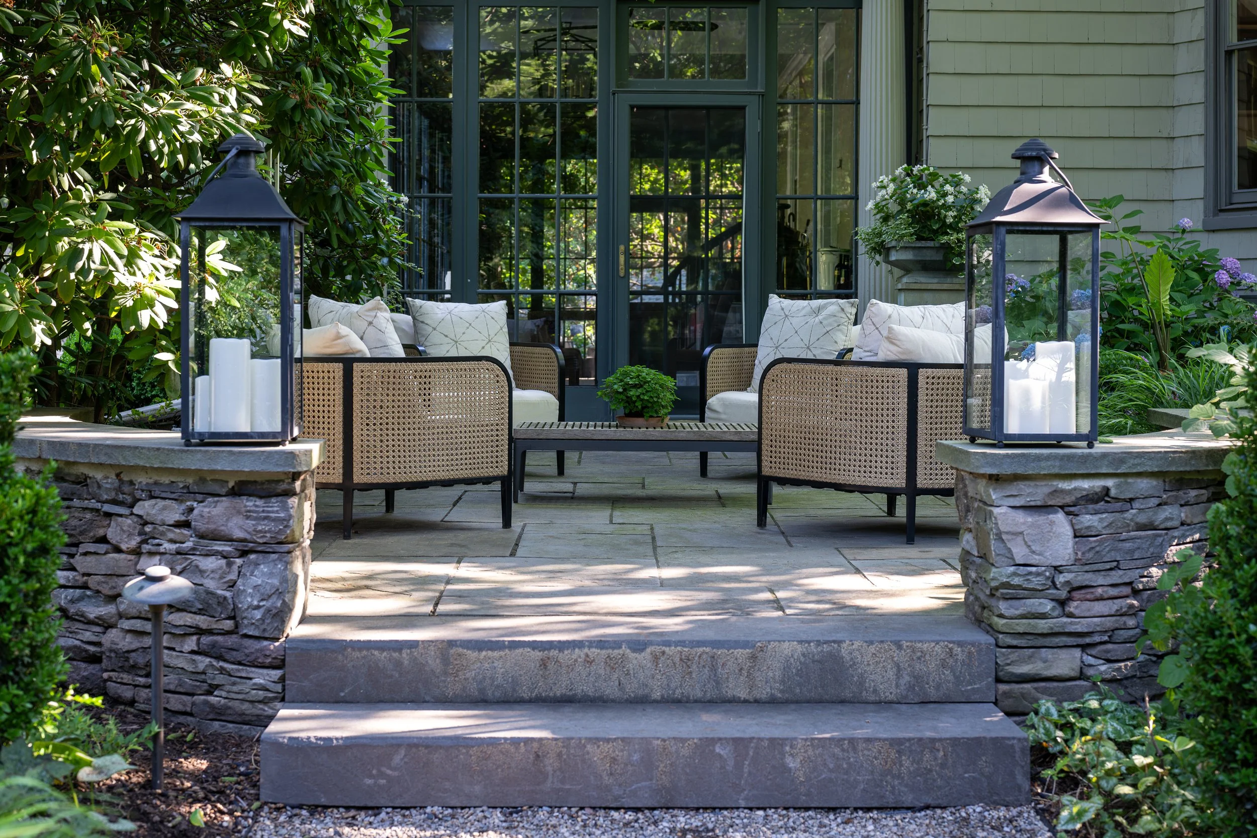 A cozy outdoor patio with stone steps leading up to a seating area, featuring a wooden table, cushioned chairs, plants, and lanterns, surrounded by greenery and trees.