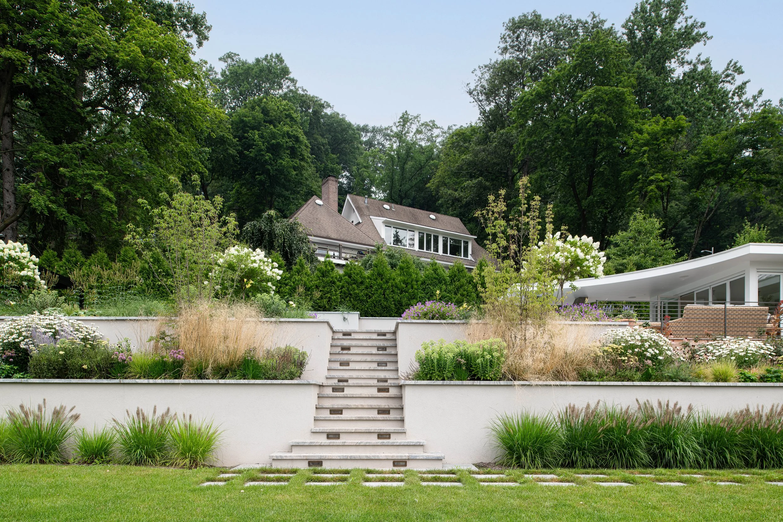 A modern backyard garden with tiered retaining walls, lush greenery, flowering plants, and a house with large windows in the background.